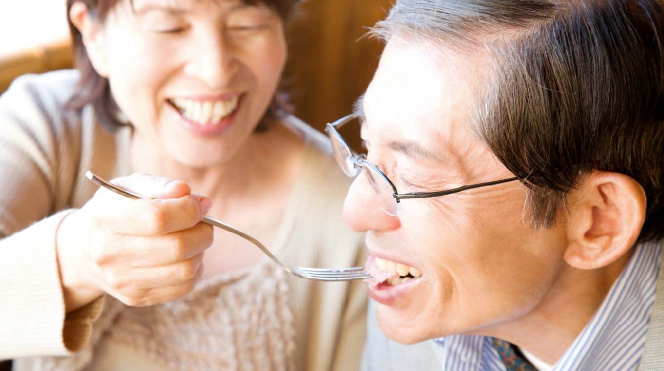 高齢の夫婦の写真。妻がフォークに食事を乗せて夫の口元へ運んでいる。夫は白い歯を見せて嬉しそうに食べている。